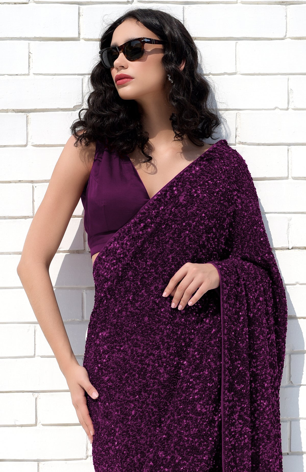 A woman in a purple sequin saree and matching blouse poses against a white brick wall.