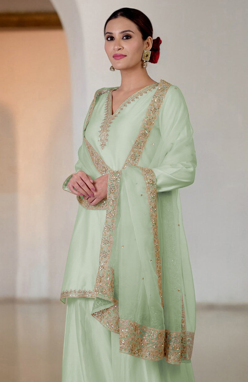A woman models a sage green sharara set with gold embroidery and a matching dupatta.