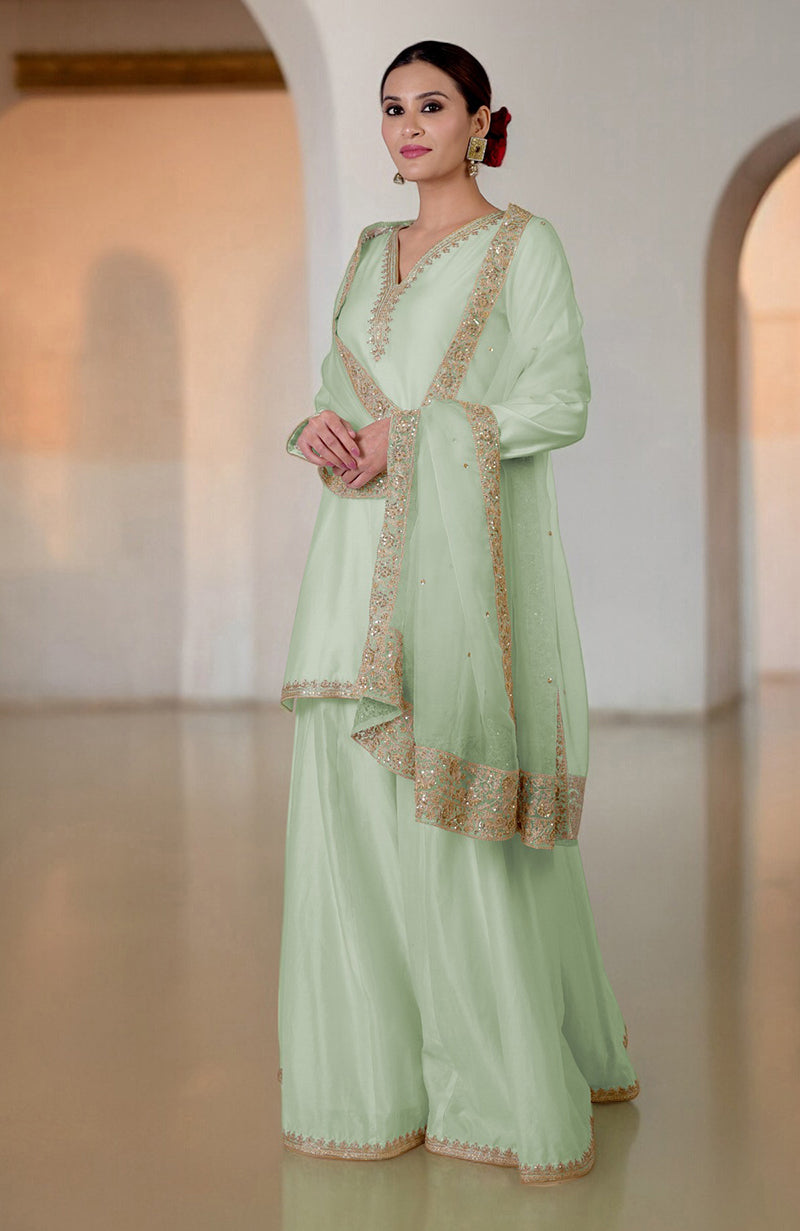 A woman models a sage green sharara set with gold embroidery and a matching dupatta.