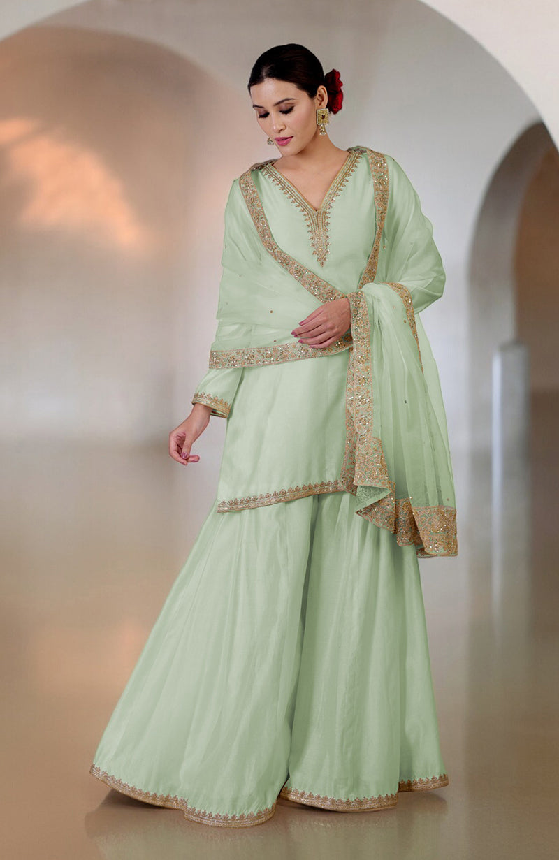 A woman models a light green sharara set with gold embroidery and a matching dupatta.