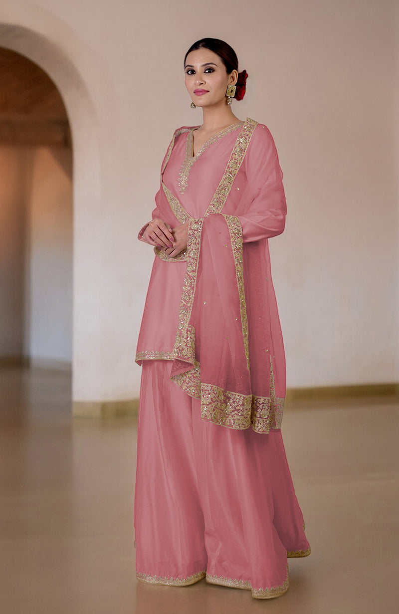 A woman models a pink sharara set with gold embroidery and a matching dupatta.