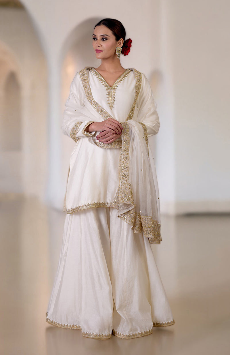 A woman models a white sharara set with gold embroidery and a matching dupatta.