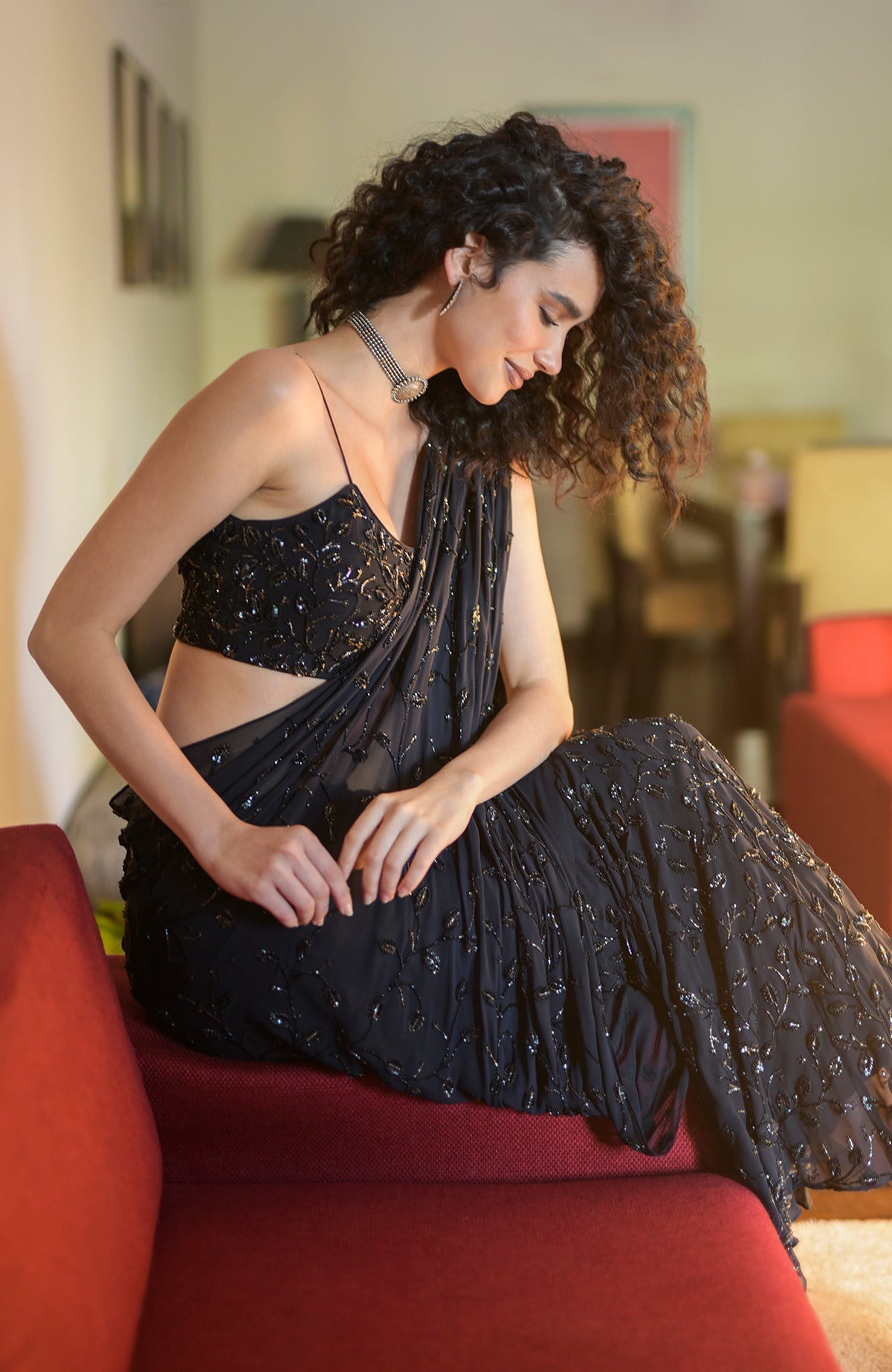 A woman in a black hand-beaded embroidered draped gown saree sits on a red couch.