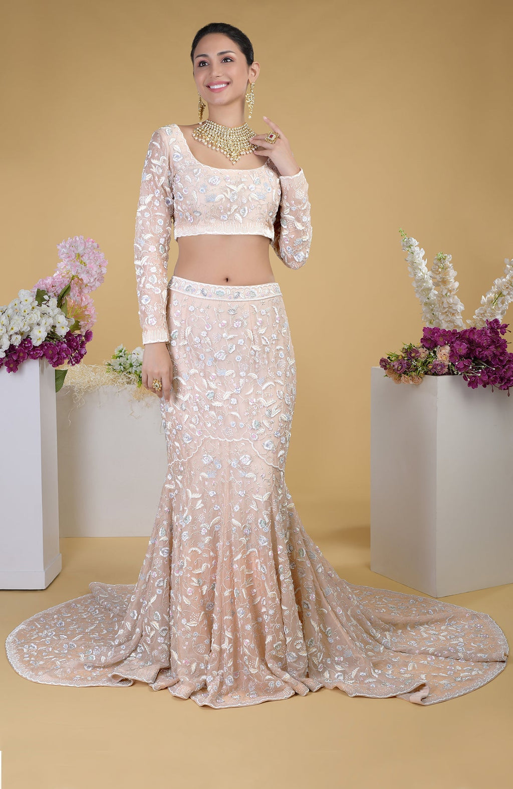 A woman models a peach embroidered crop top and matching mermaid skirt with a long train.
