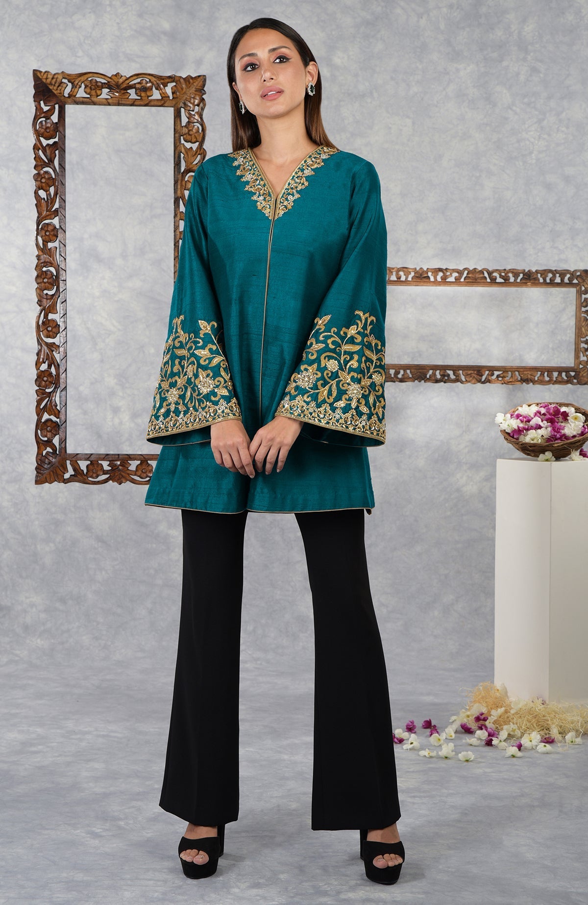 A woman models a teal raw silk jacket with gold embroidery and black flared pants.