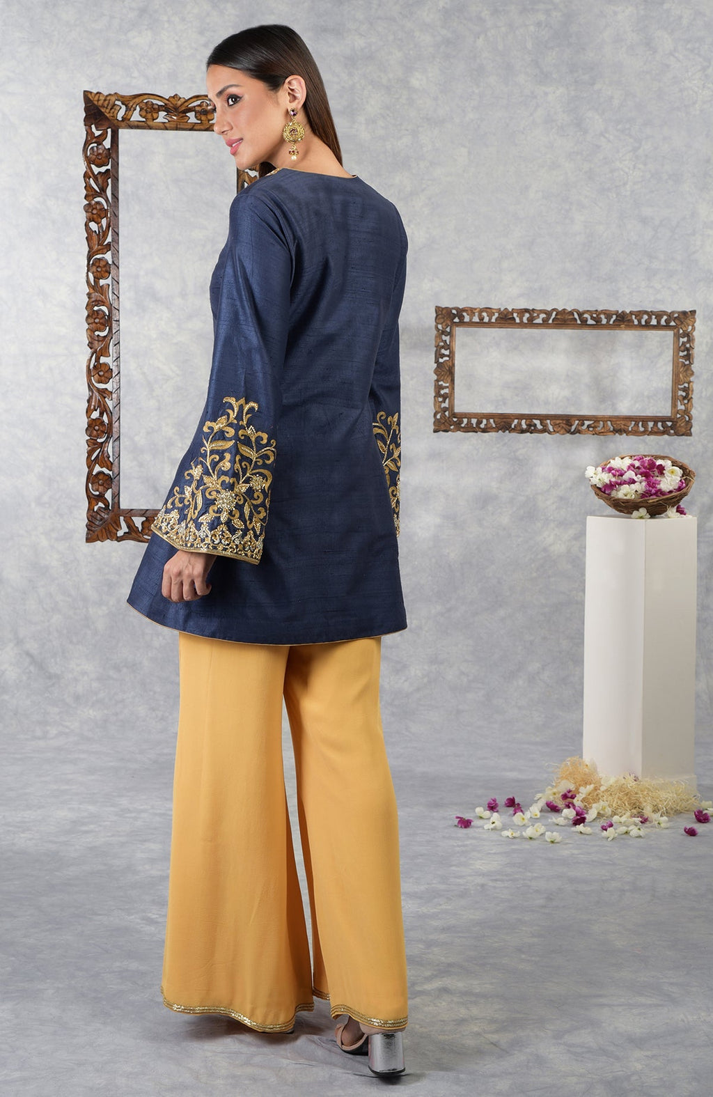 A woman models a navy blue raw silk jacket with gold embroidery and honey yellow palazzo pants.