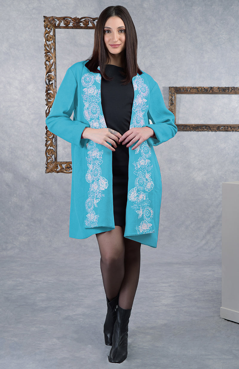 A woman models a teal open-front jacket with white floral embroidery over a black dress.
