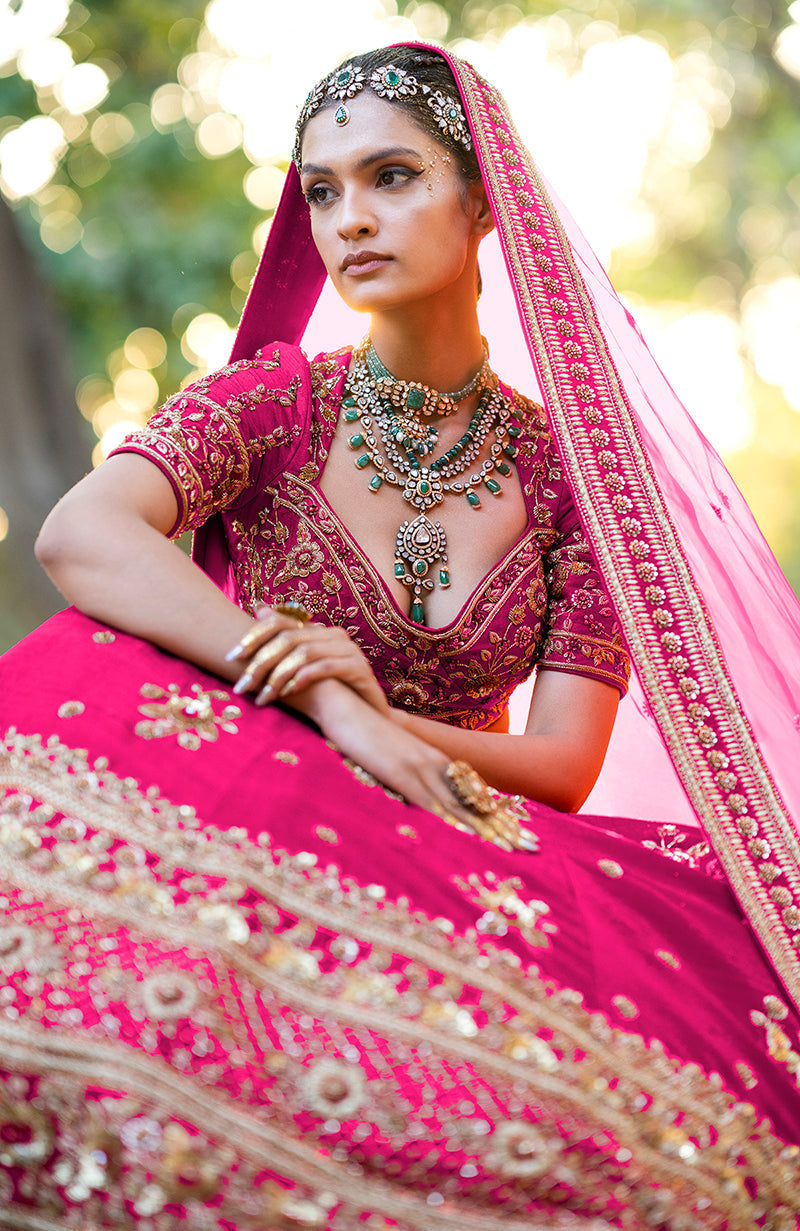 A woman models a hot pink lehenga set with intricate gold embroidery and emerald jewelry.