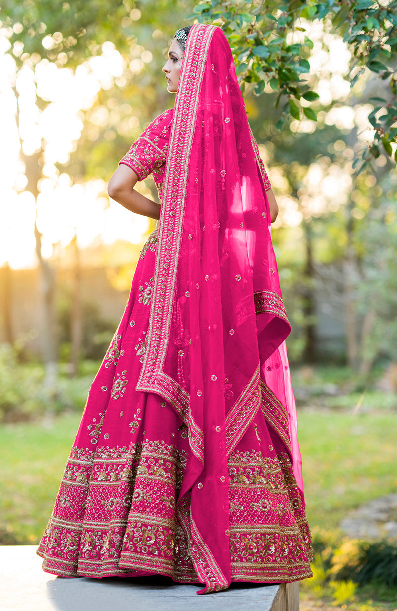 A woman models a hot pink lehenga set with gold embroidery and a sheer dupatta.