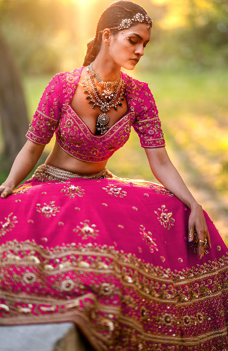 A model wears a fuchsia lehenga set with intricate gold embroidery and jewelry.