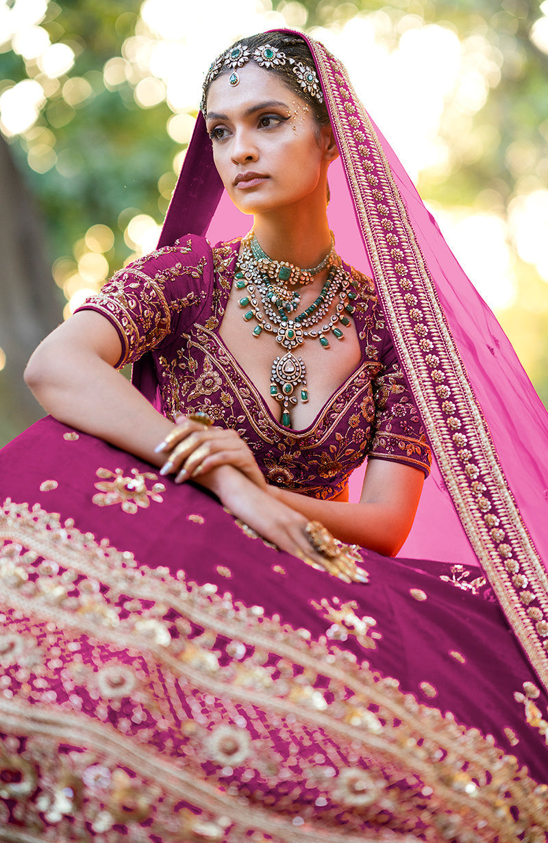 A woman models a purple wine lehenga set with gold embroidery and emerald jewelry.
