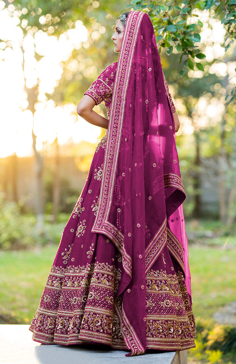 A model in a purple wine lehenga set with gold embroidery stands outdoors.