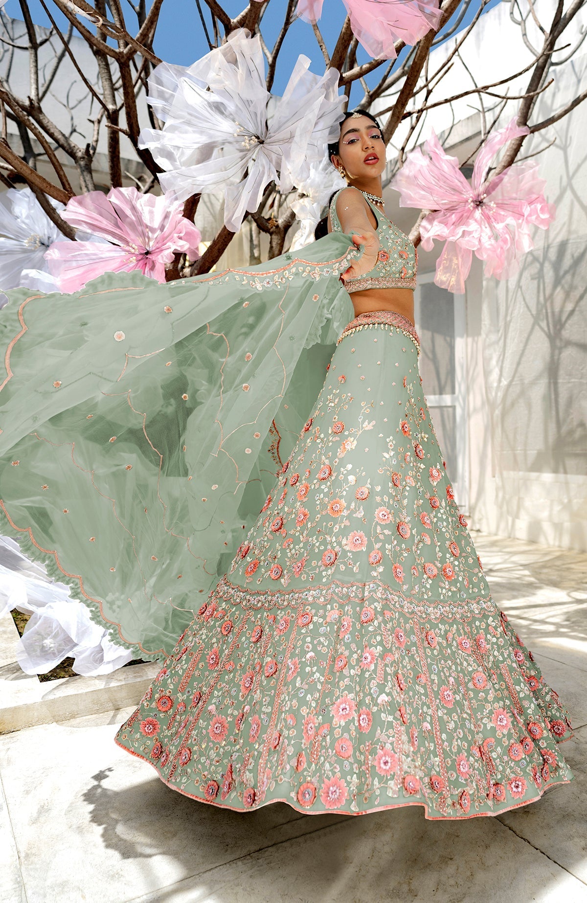 A model poses in a sage green lehenga with pink floral embroidery and a sheer dupatta.