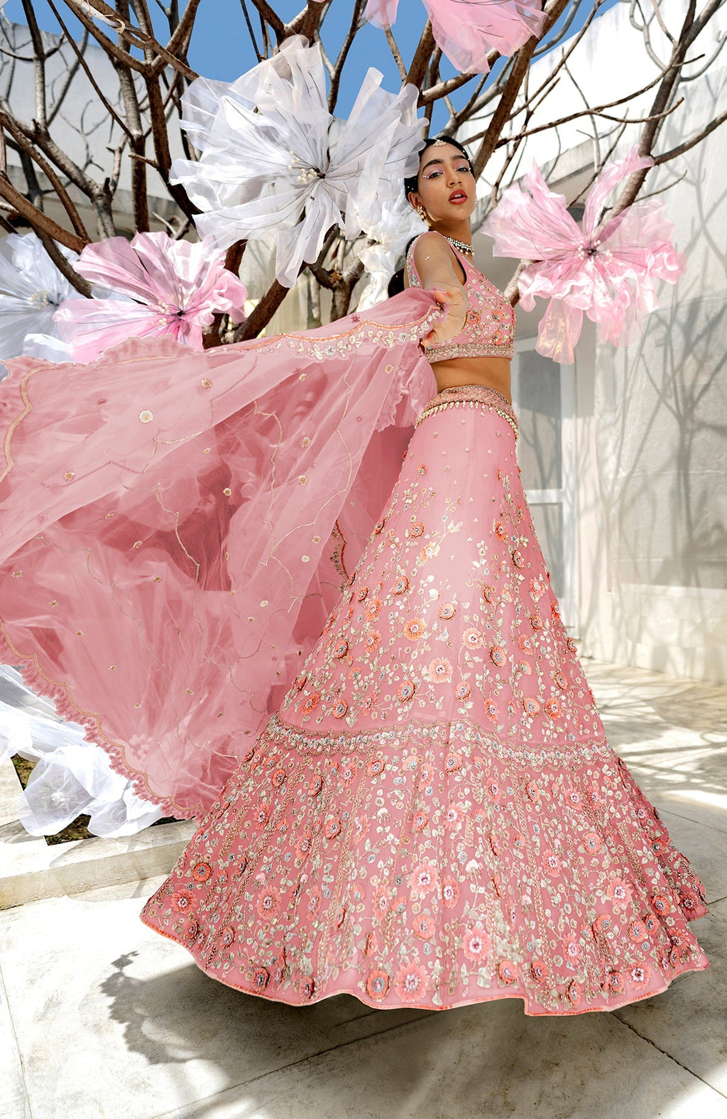 A model poses in a pink lehenga set with intricate floral embroidery and a sheer dupatta.
