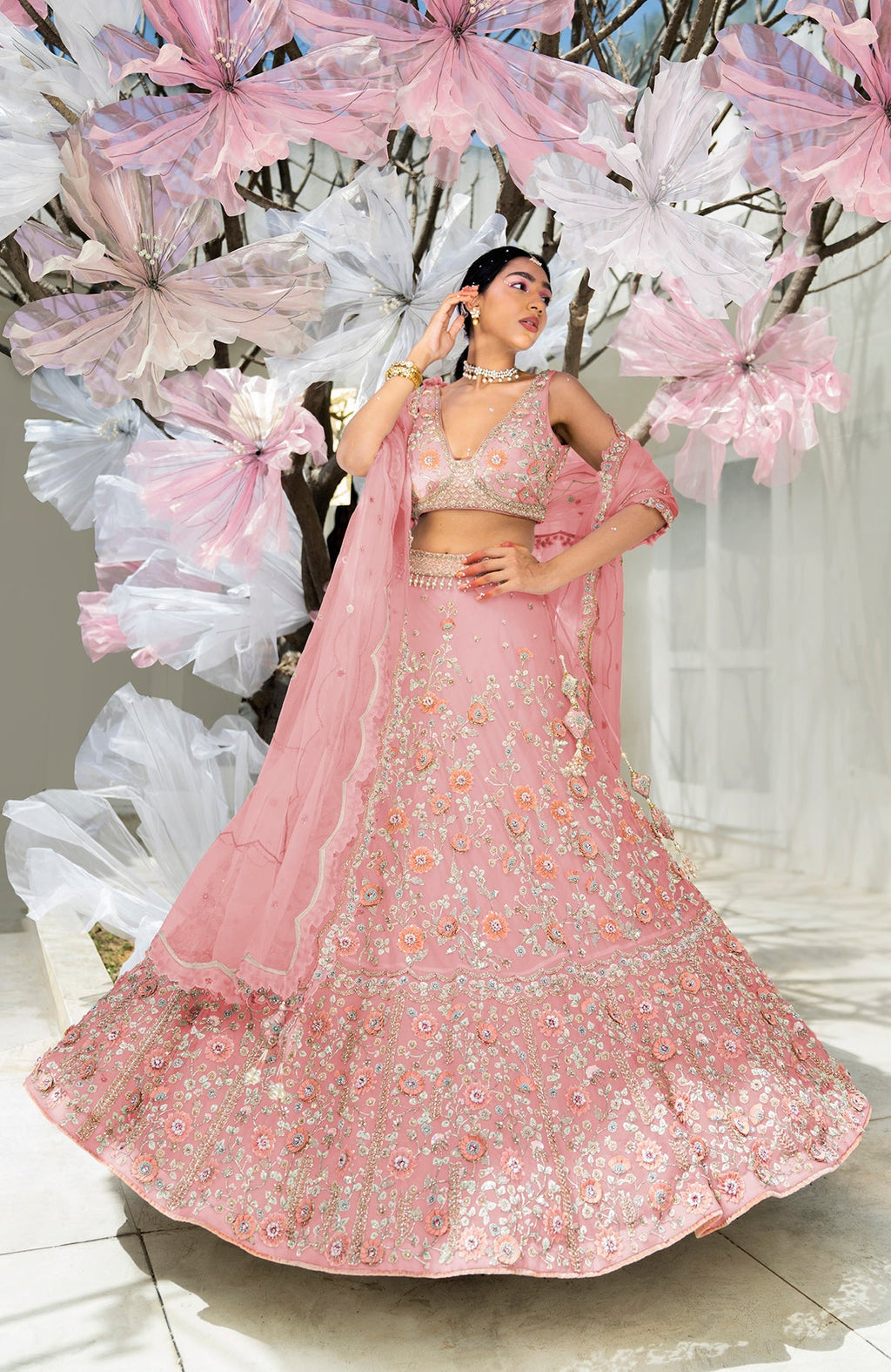 A woman in a pink floral embroidered lehenga set poses against a floral backdrop.