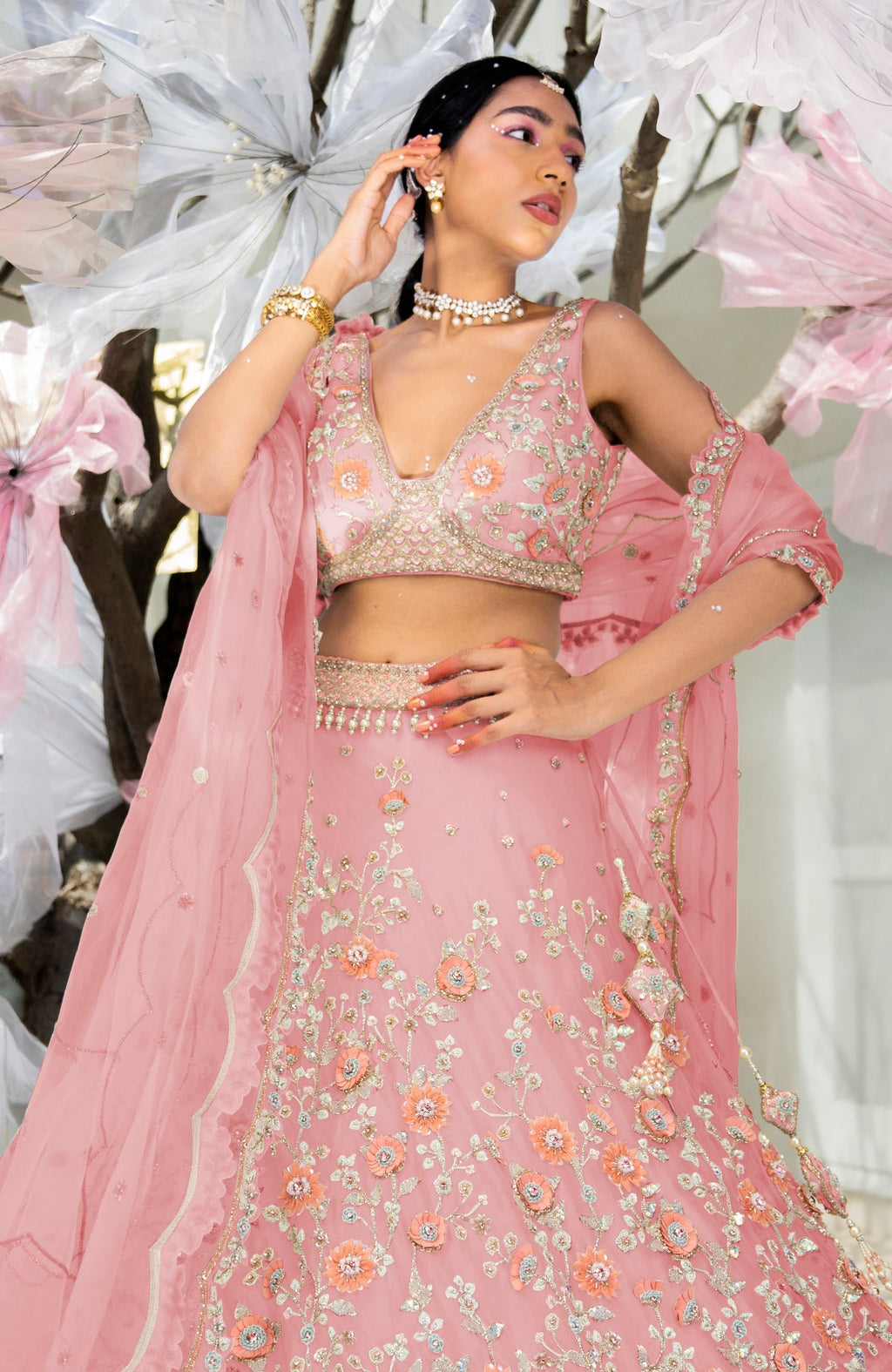 A woman models a pink lehenga set with intricate floral embroidery and jewelry.