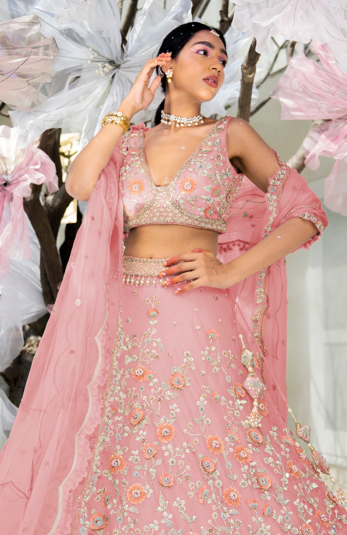 A woman models a pink lehenga set with intricate floral embroidery and jewelry.
