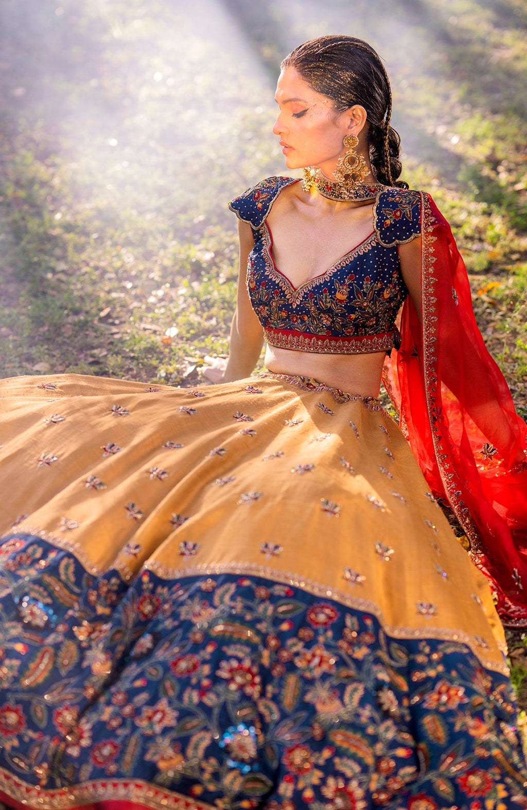 A model sits wearing a mustard and blue embroidered lehenga with a red dupatta.