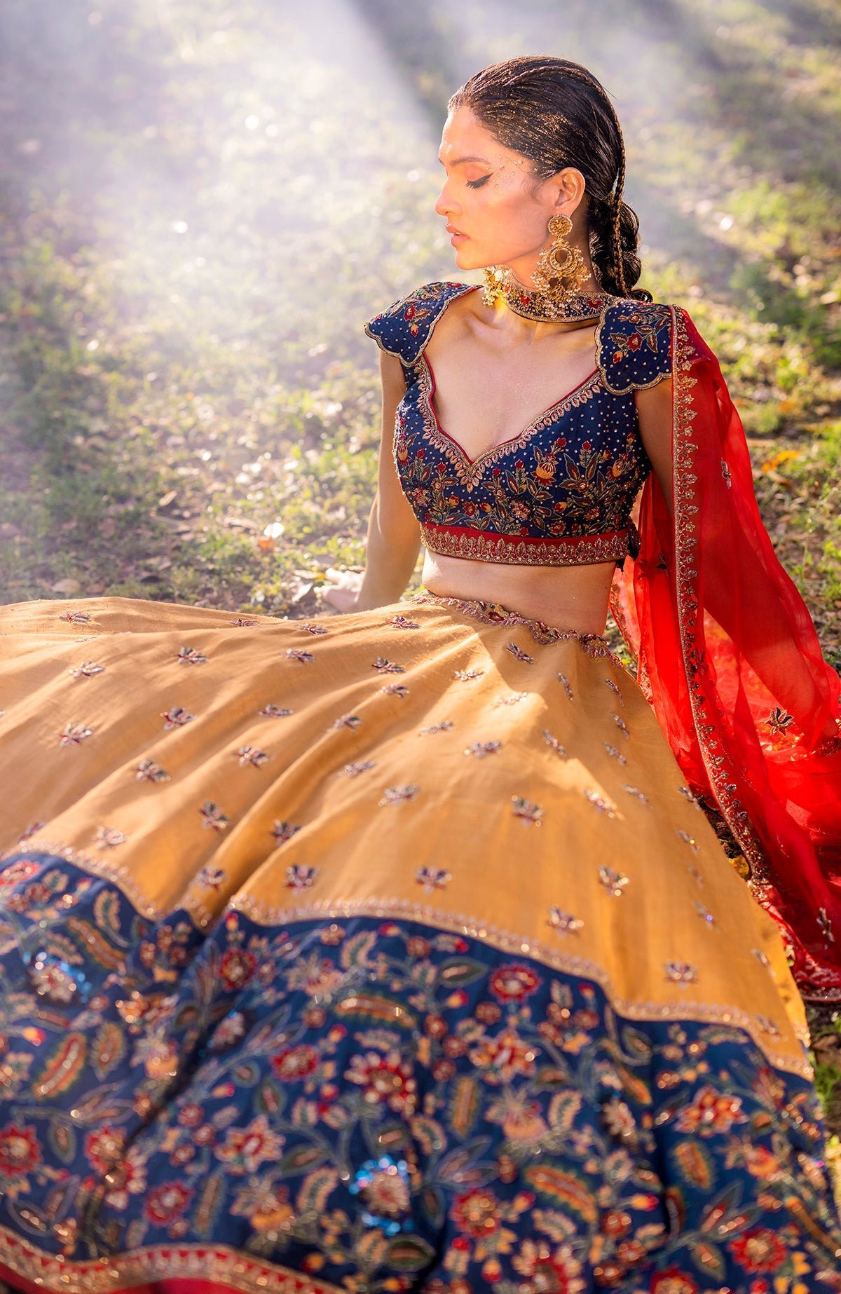 A model sits wearing a mustard and blue embroidered lehenga with a red dupatta.
