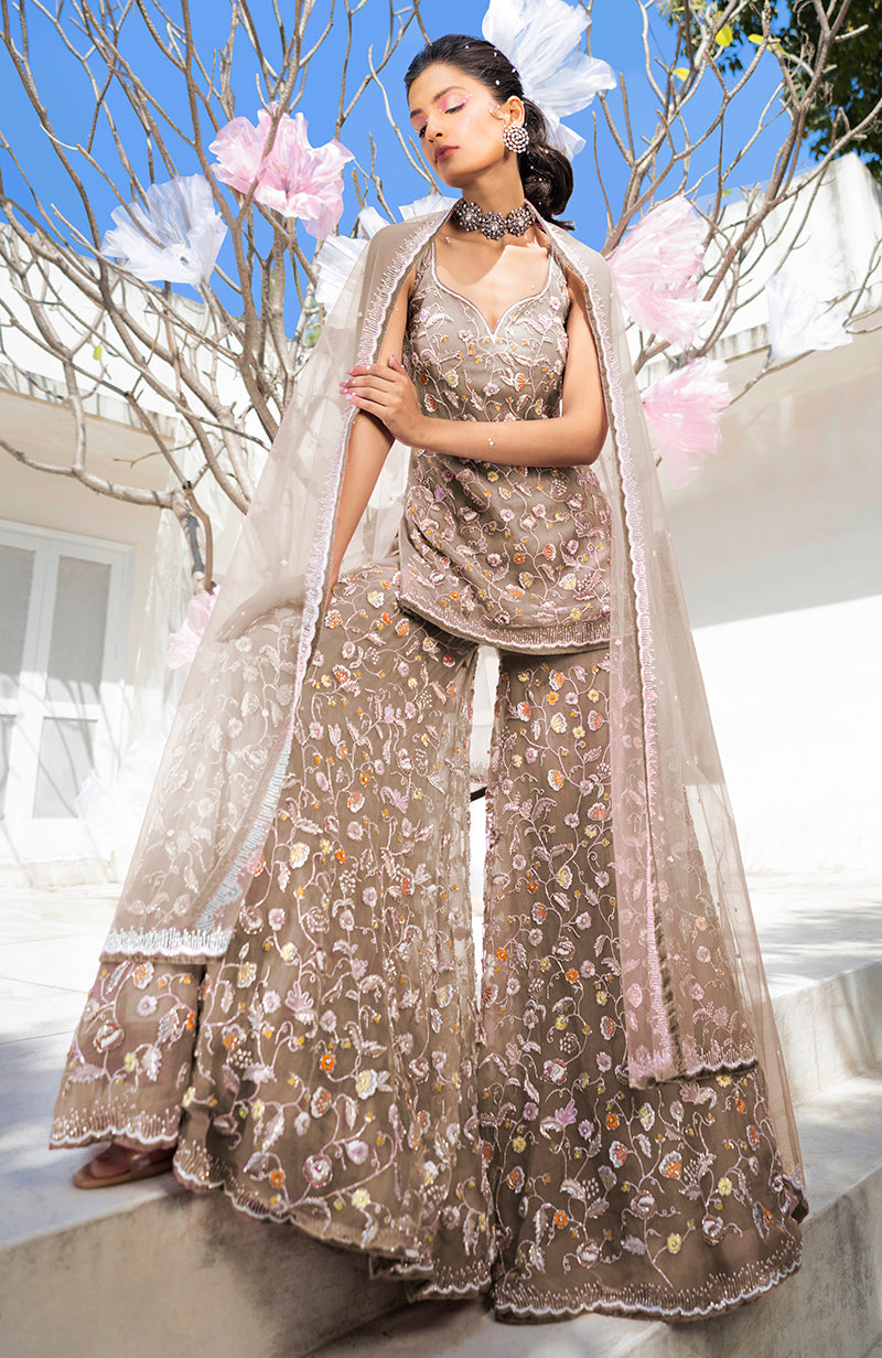 A woman models a violet sharara set with intricate floral embroidery and a sheer dupatta.
