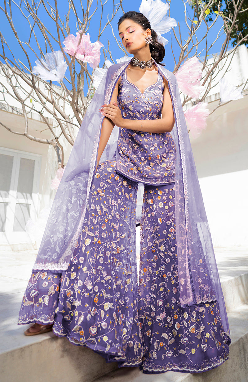 A woman models a violet sharara set with floral embroidery and a sheer dupatta.