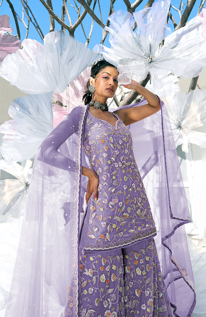 A woman in a violet sharara set with floral embroidery poses against a floral backdrop.