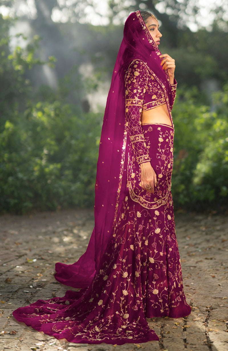 A woman in a peacock pink lehenga set with gold embroidery stands outdoors.
