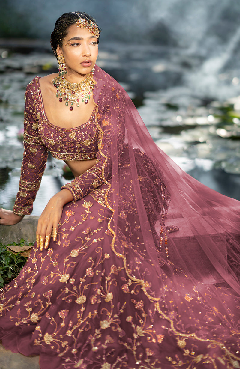 A woman in a rose wine lehenga set with gold embroidery sits by water.