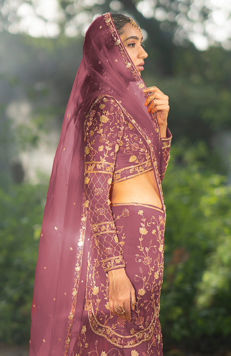 A woman models a rose wine lehenga set with gold embroidery and a sheer dupatta.