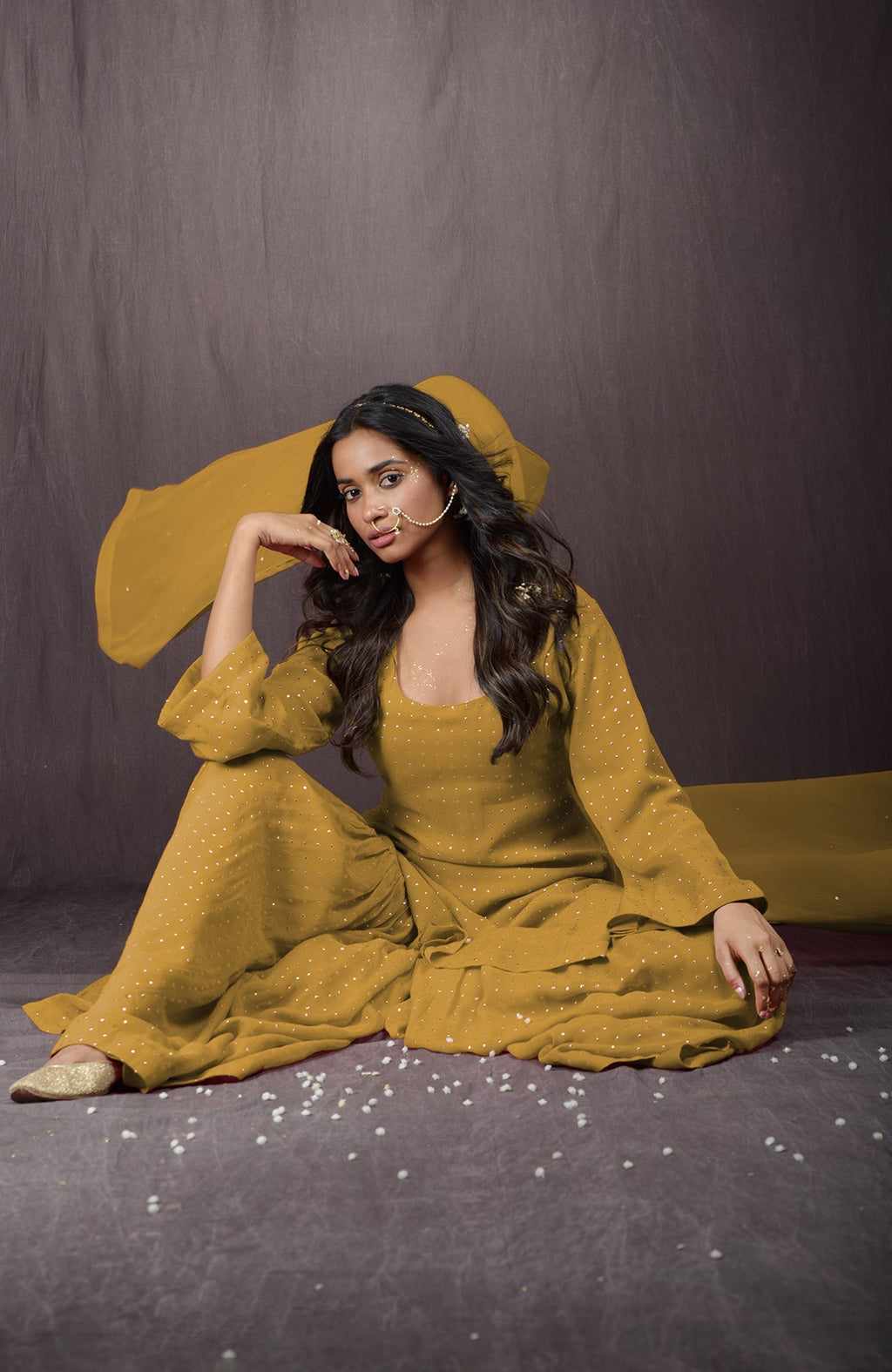 A woman in a mustard yellow sharara set with gold embellishments sits on a dark background.