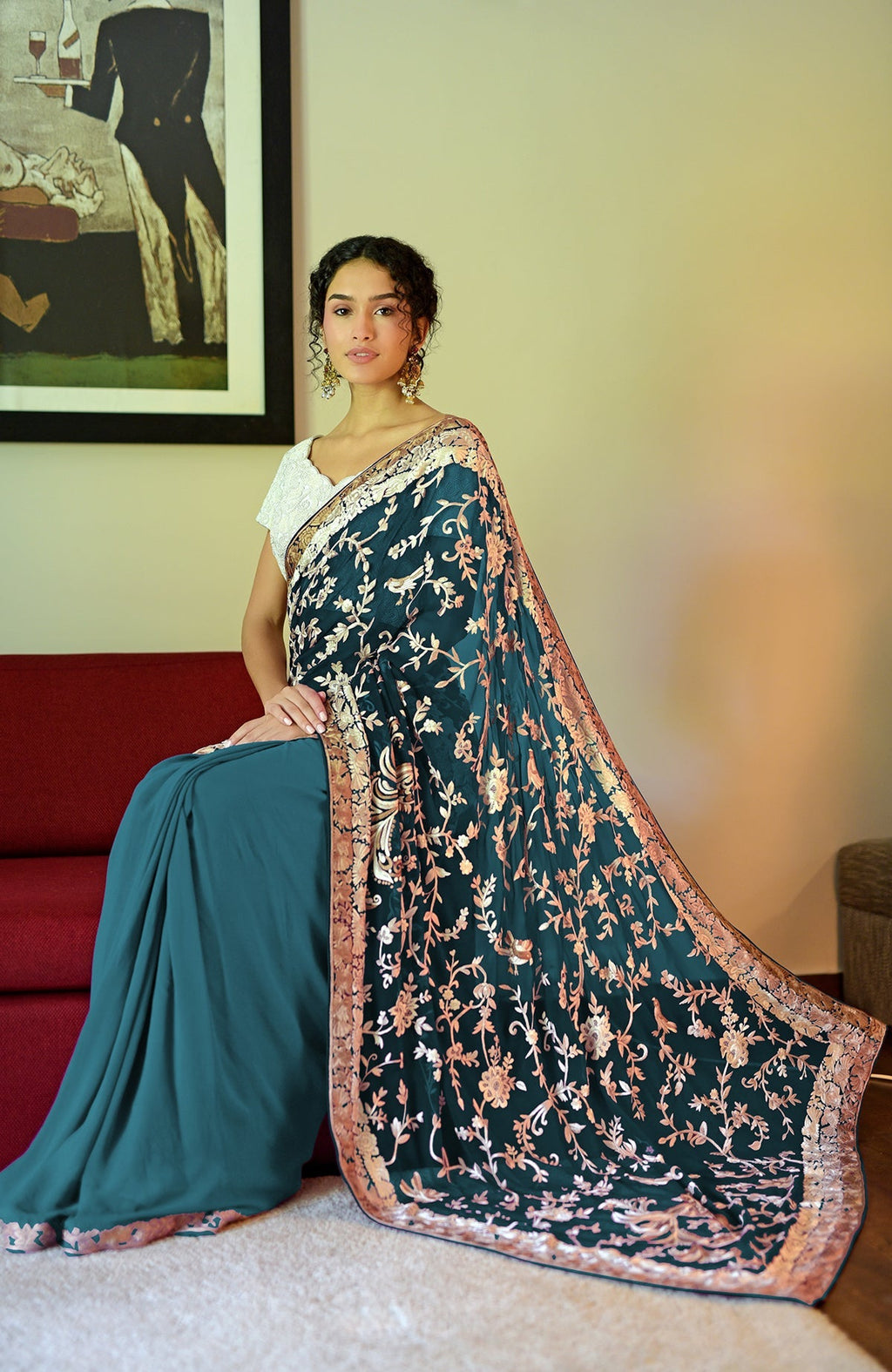 A woman in a peacock blue-teal Parsi Gara hand-embroidered saree sits on a red couch.