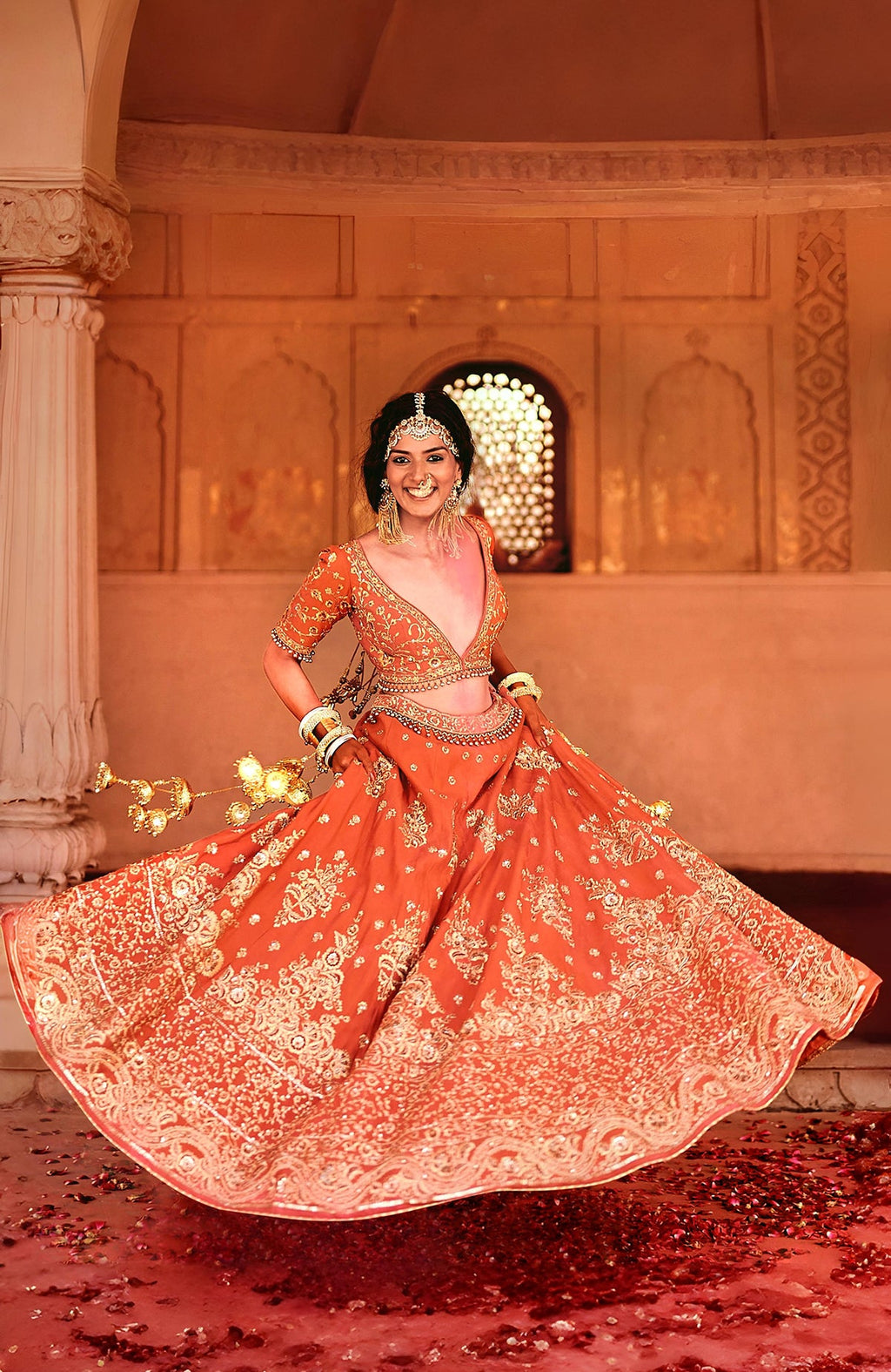 A smiling woman in a red lehenga with gold embroidery twirls, holding string lights.