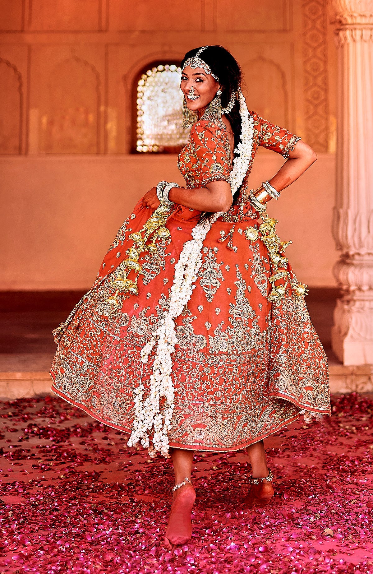 A woman in a red lehenga with gold embroidery and white floral garlands smiles over her shoulder.