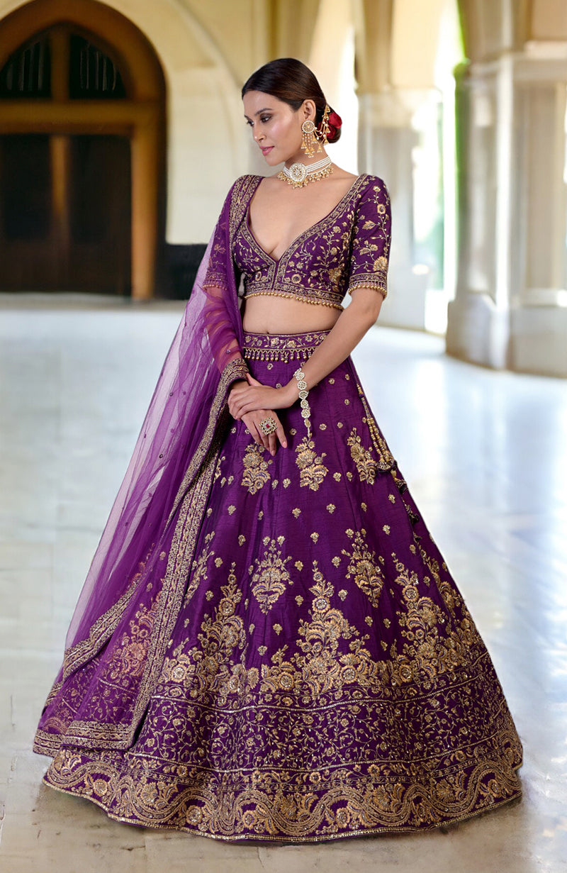 A woman models a plum lehenga set with gold embroidery and a sheer dupatta.