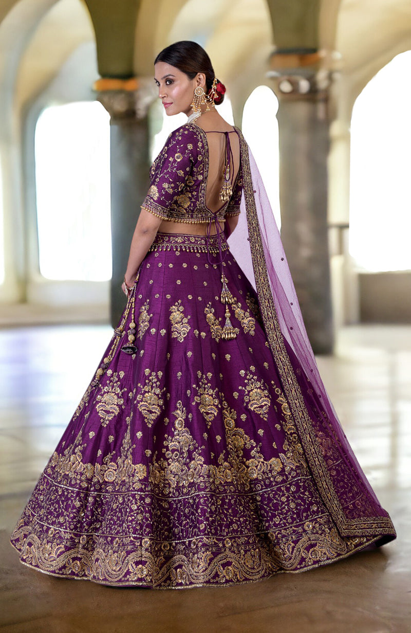 A woman models a plum lehenga set with intricate gold embroidery and an open back.