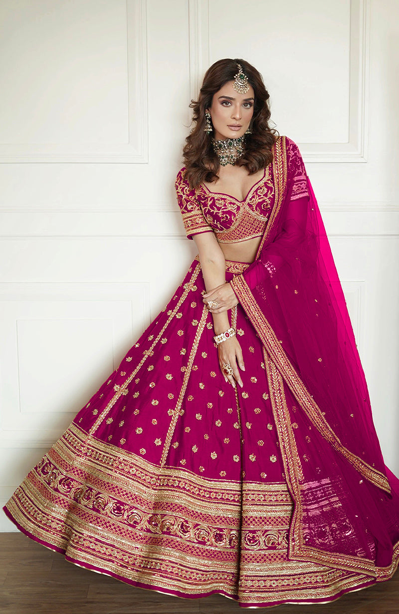 A woman models a fuchsia fedora lehenga set with gold embroidery and a matching dupatta.