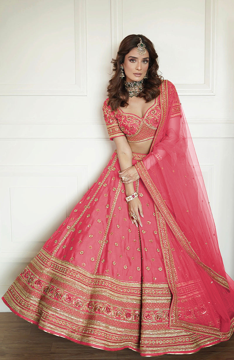 A woman models a flamingo pink lehenga set with gold embroidery and a sheer dupatta.