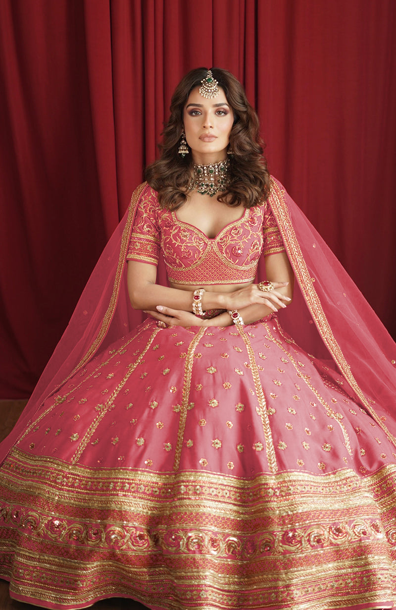 A woman models a flamingo pink lehenga set with gold embroidery and a sheer dupatta.