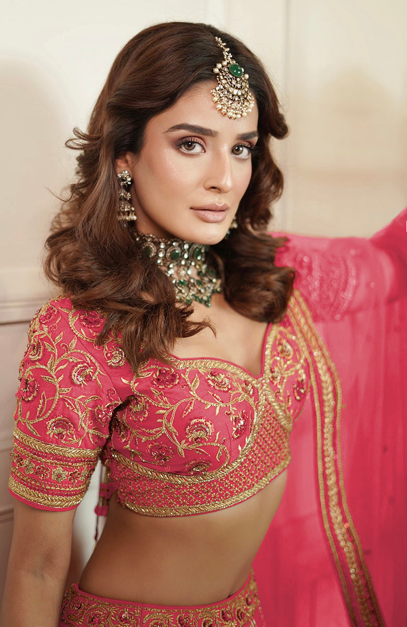 A woman in a flamingo pink lehenga set with gold embroidery and green jewelry.