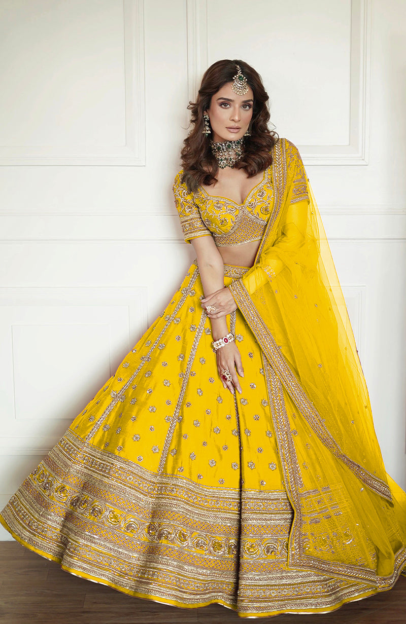 A woman models a yellow lehenga set with intricate gold embroidery and a sheer dupatta.