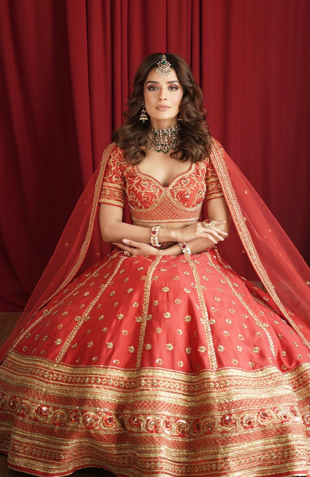 A woman models a scarlet red lehenga set with gold embroidery and a sheer dupatta.