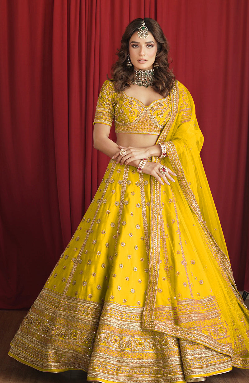A woman models a bright yellow lehenga set with intricate gold embroidery.