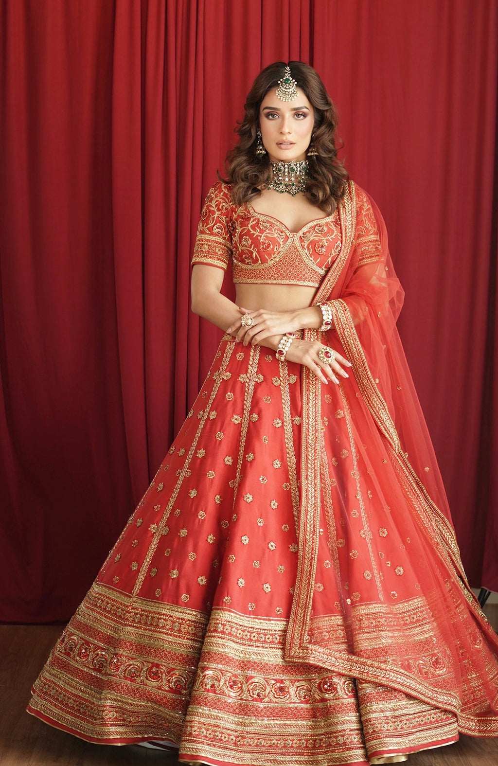A woman models a scarlet red lehenga set with gold embroidery and green jewelry.