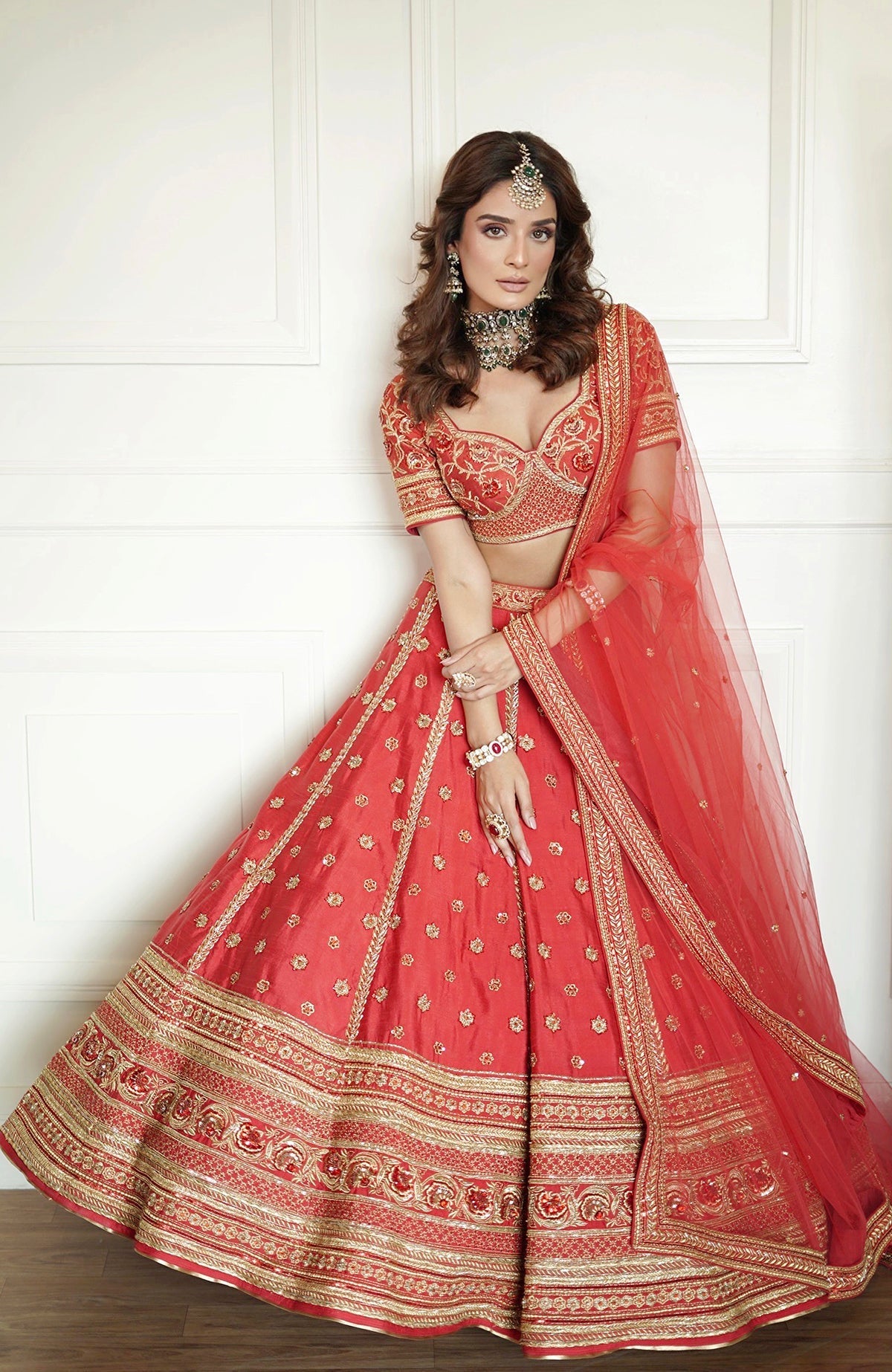 A woman models a red lehenga set with gold embroidery and traditional jewelry.