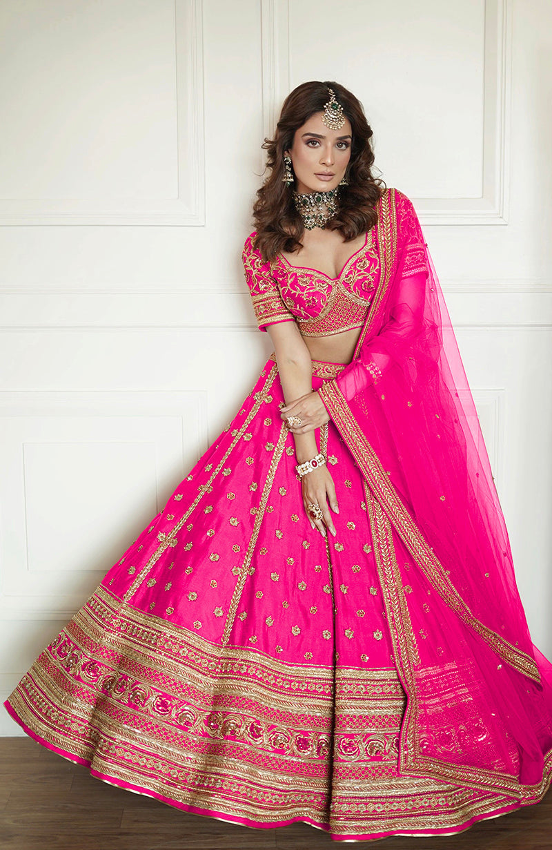 A woman models a flamingo pink lehenga set with gold embroidery and a sheer dupatta.