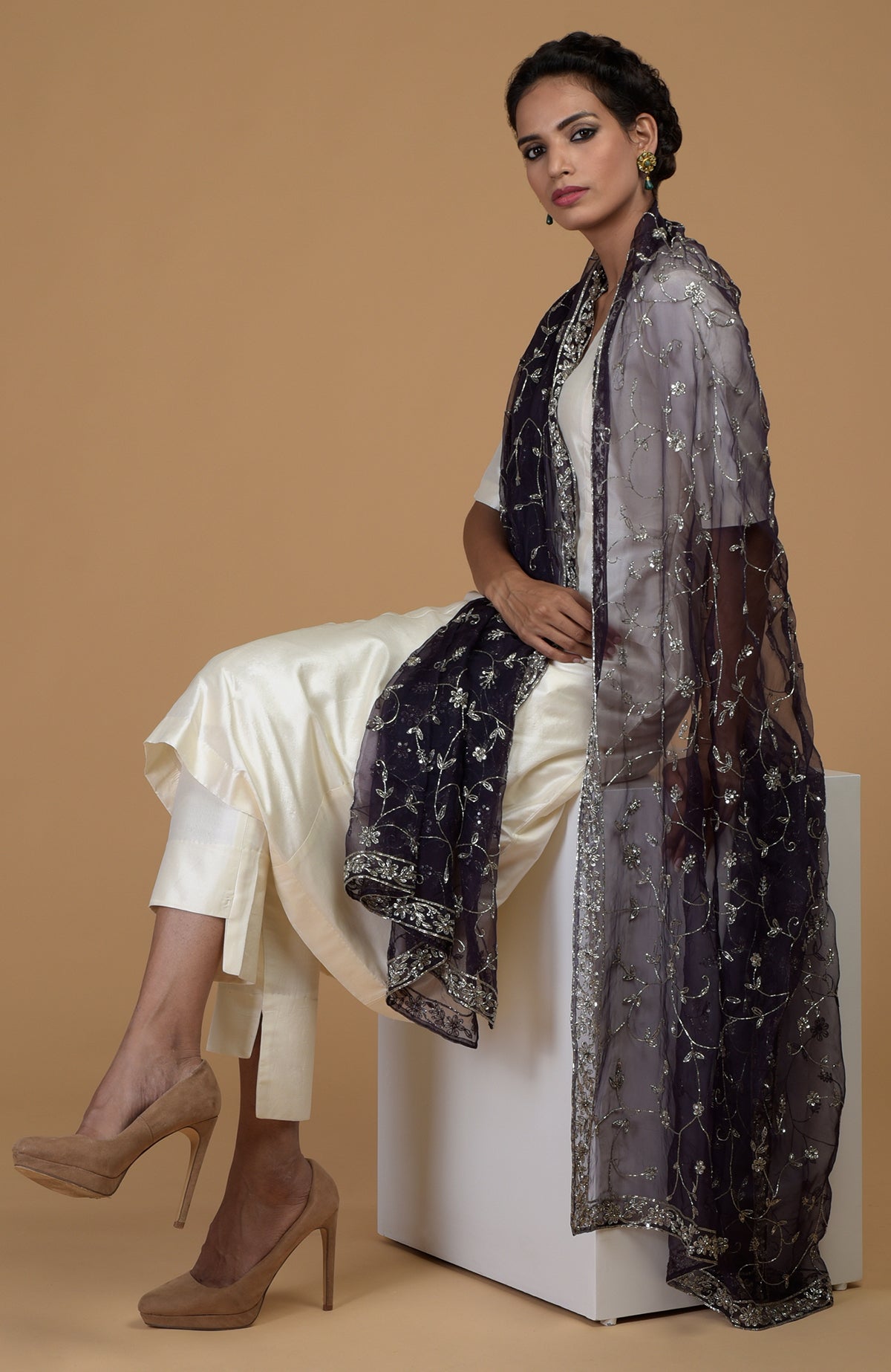Woman in an ivory suit with an eclipse blue embroidered dupatta, sitting on a white block.