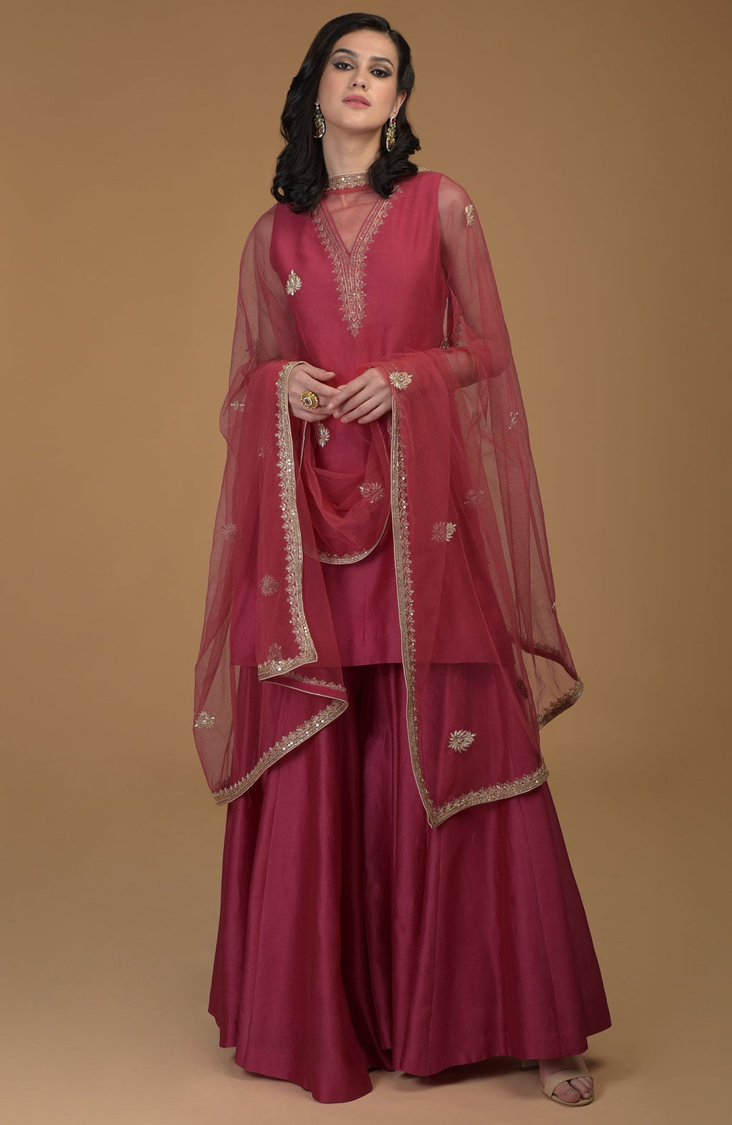 A woman models a ruby red gharara suit with gold zardozi embroidery.