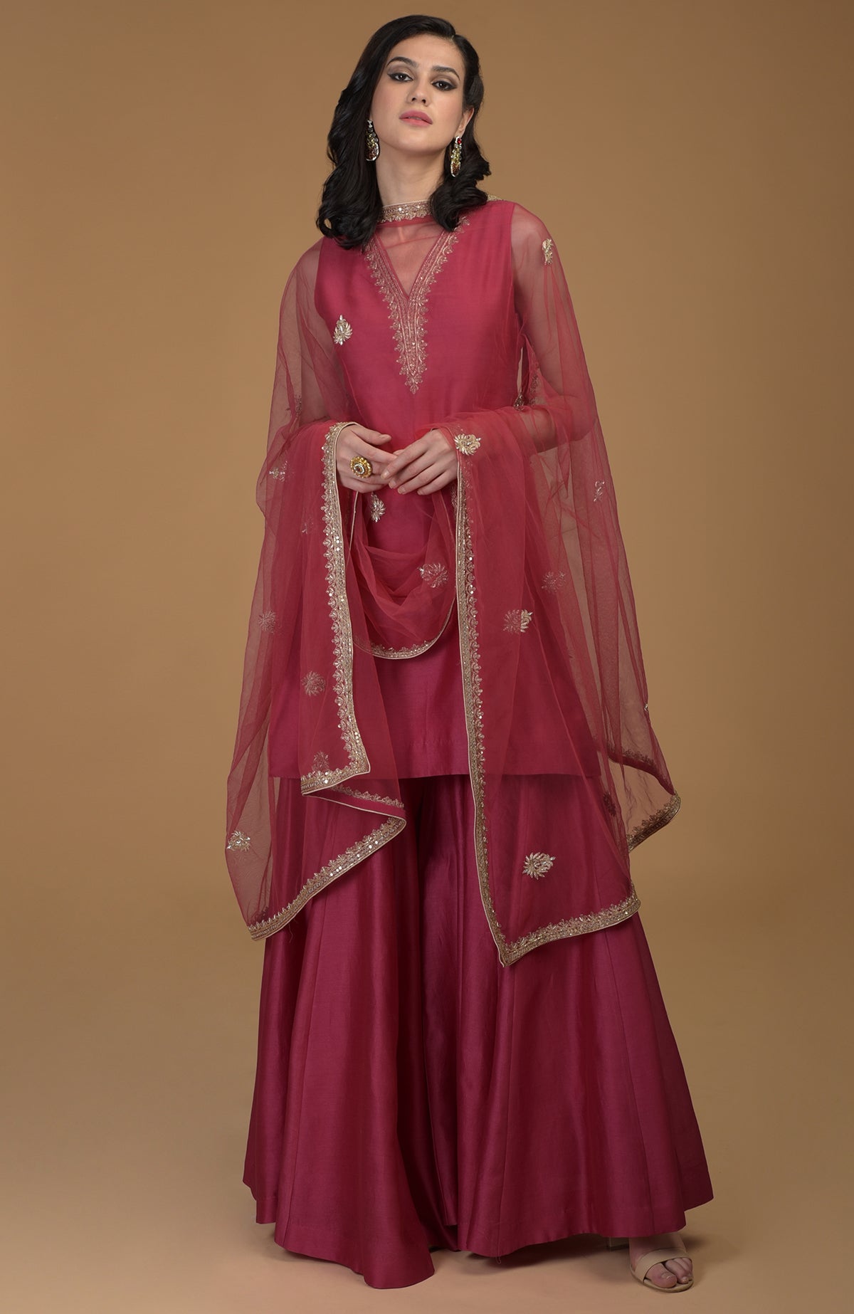 A woman models a ruby red gharara suit with gold zardozi embroidery.