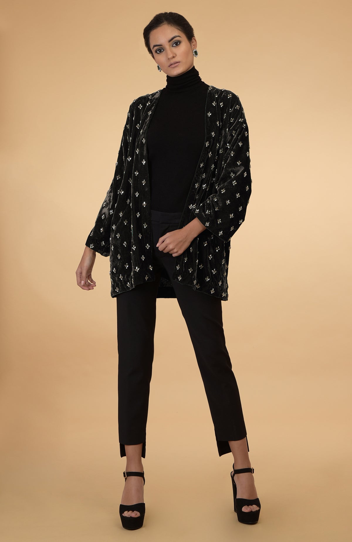 A woman models a dark olive crystal hand-embroidered silk velvet jacket over a black turtleneck and pants.