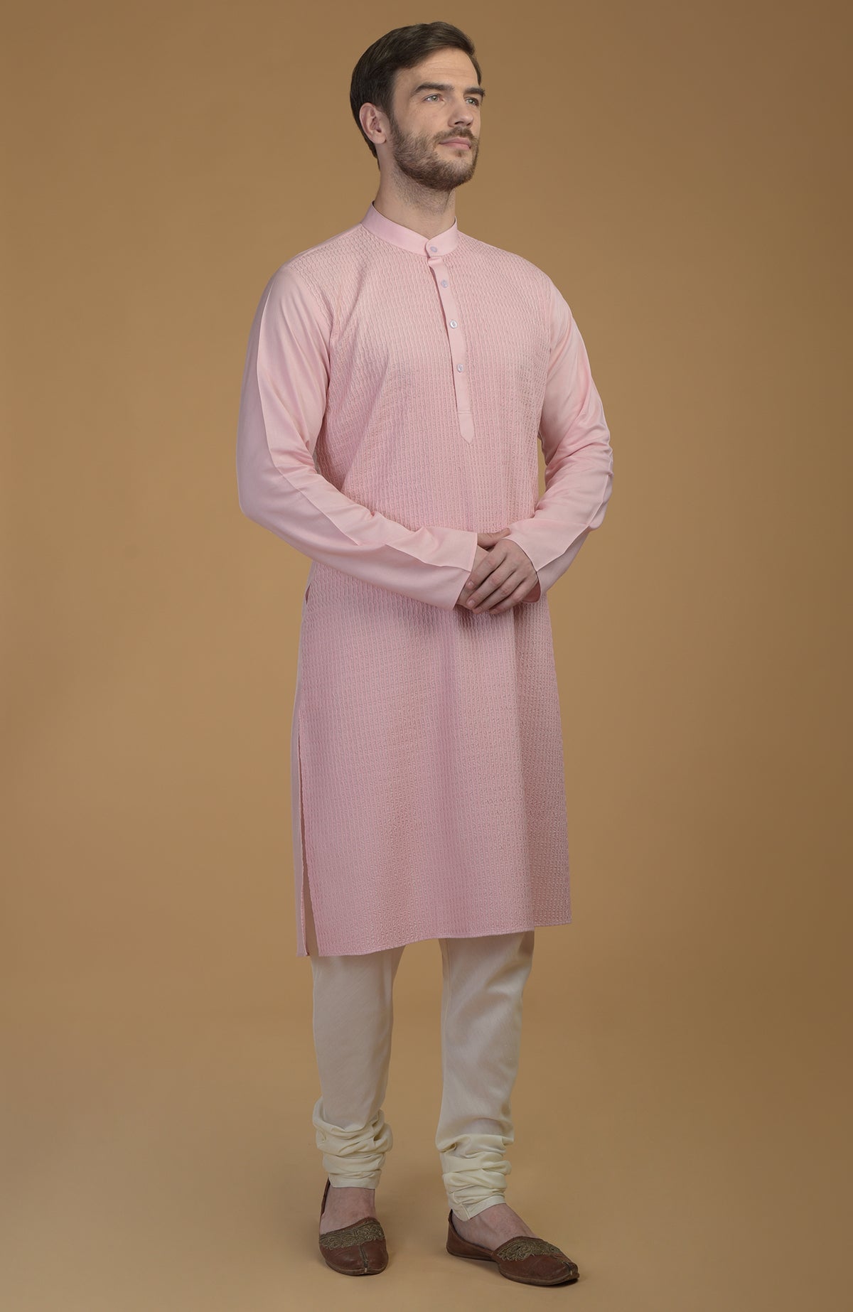A man stands wearing a pale pink kurta with pintuck detail and ivory churidars.